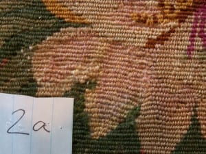 Conservation documentation photo labeled "2a" showing the same floral needlework area after treatment, with the previously damaged hole now stabilized and re-woven with blended pink and tan wool stitching matching the surrounding pattern.