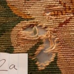 Conservation documentation photo labeled "2a" showing a jagged hole with loose unraveled wool threads in a floral needlework chair seat, with green, tan, and pink surrounding stitching intact.