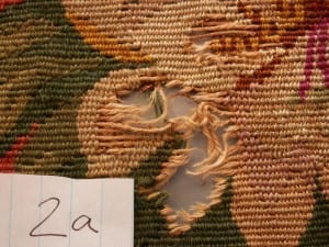 Conservation documentation photo labeled "2a" showing a jagged hole with loose unraveled wool threads in a floral needlework chair seat, with green, tan, and pink surrounding stitching intact.