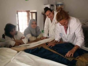 Julia Brennan in a white lab coat shows three Tunisian colleagues how to handle a dark blue velvet and gold embroidered garment on a worktable.