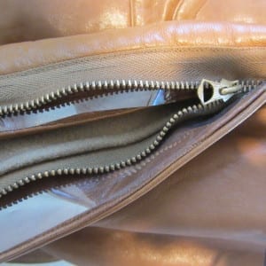 A tan leather flight suit showing a cleaned brass zipper with pull tab, now free of corrosion, with the open zipper revealing the brown lining and interior pockets of the garment.