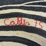 A fiber tapestry corner showing the signature "Calder 75" stitched in red cord against a black and cream striped woven background.