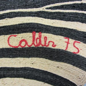 A fiber tapestry corner showing the signature "Calder 75" stitched in red cord against a black and cream striped woven background.
