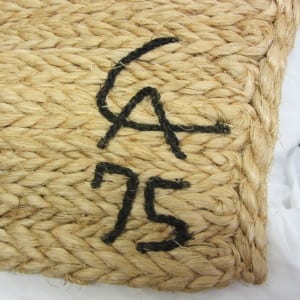 Corner of a natural fiber tapestry showing a hand-stitched artist's monogram and the date "75" in black cord.