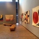 A museum gallery with five large abstract fiber tapestries featuring bold spiral, circular, and geometric designs in red, black, yellow, and cream, hung along gray walls.