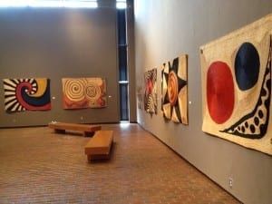 A museum gallery with five large abstract fiber tapestries featuring bold spiral, circular, and geometric designs in red, black, yellow, and cream, hung along gray walls.