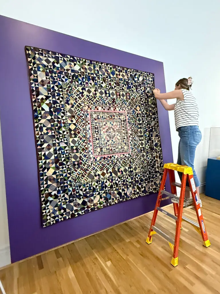 Repair of antique rug hanging on purple wall with textile worker standing on orange ladder